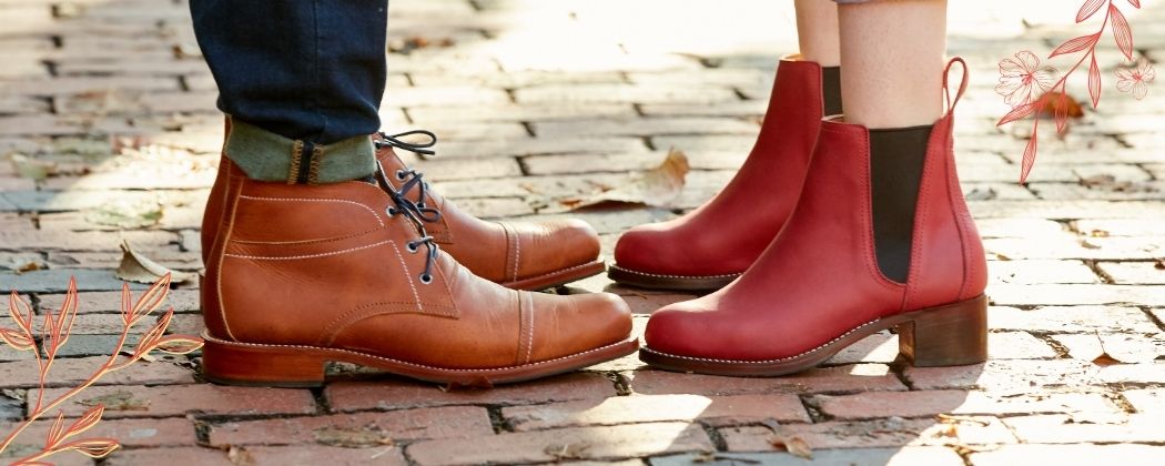 Two people standing outdoors on a brick pathway, showcasing men's brown leather lace-up boots in men's size 15 and women's red leather Chelsea boots with a block heel in a wide width.