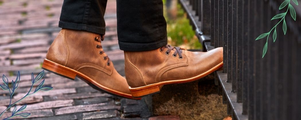 A close-up of a man wearing durable distressed brown leather lace-up boots with natural leather midsoles, stepping on a weathered stone path next to a dark metal fence for the Men's Boots Collection banner.