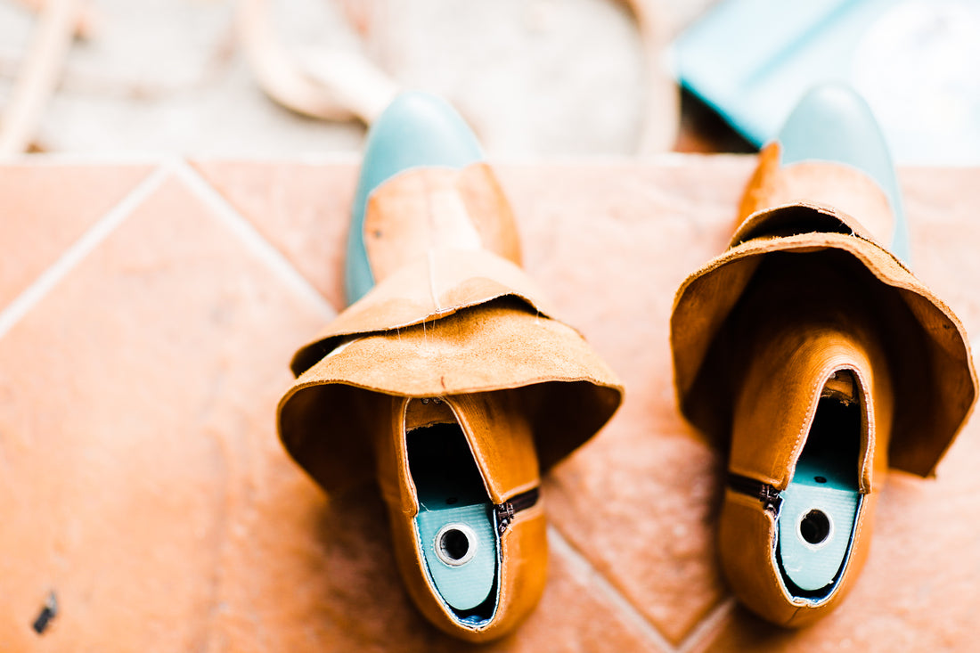 Two teal shoe uppers pulled over wooden lasts on a tile floor, mid-way through handcrafting