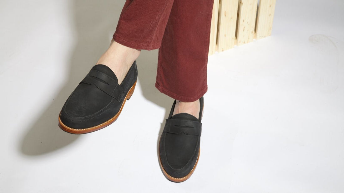 A person wearing a pair of classic women's black leather penny loafer dress shoes with tan outsoles, styled with rust-colored trousers.