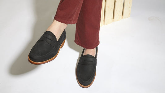 A person wearing a pair of classic women's black leather penny loafer dress shoes with tan outsoles, styled with rust-colored trousers.
