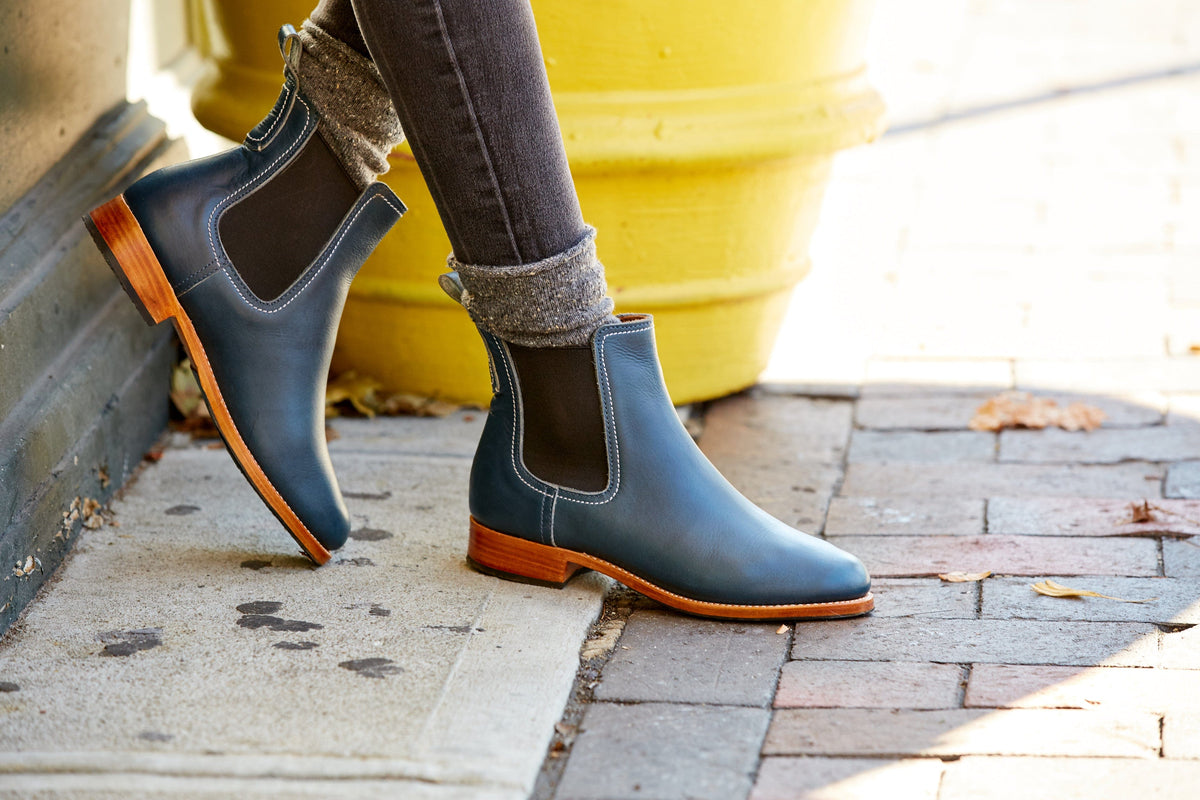 Women's dark Chelsea boots with contrast stitching and orange soles worn outdoors, available in narrow to extra wide widths