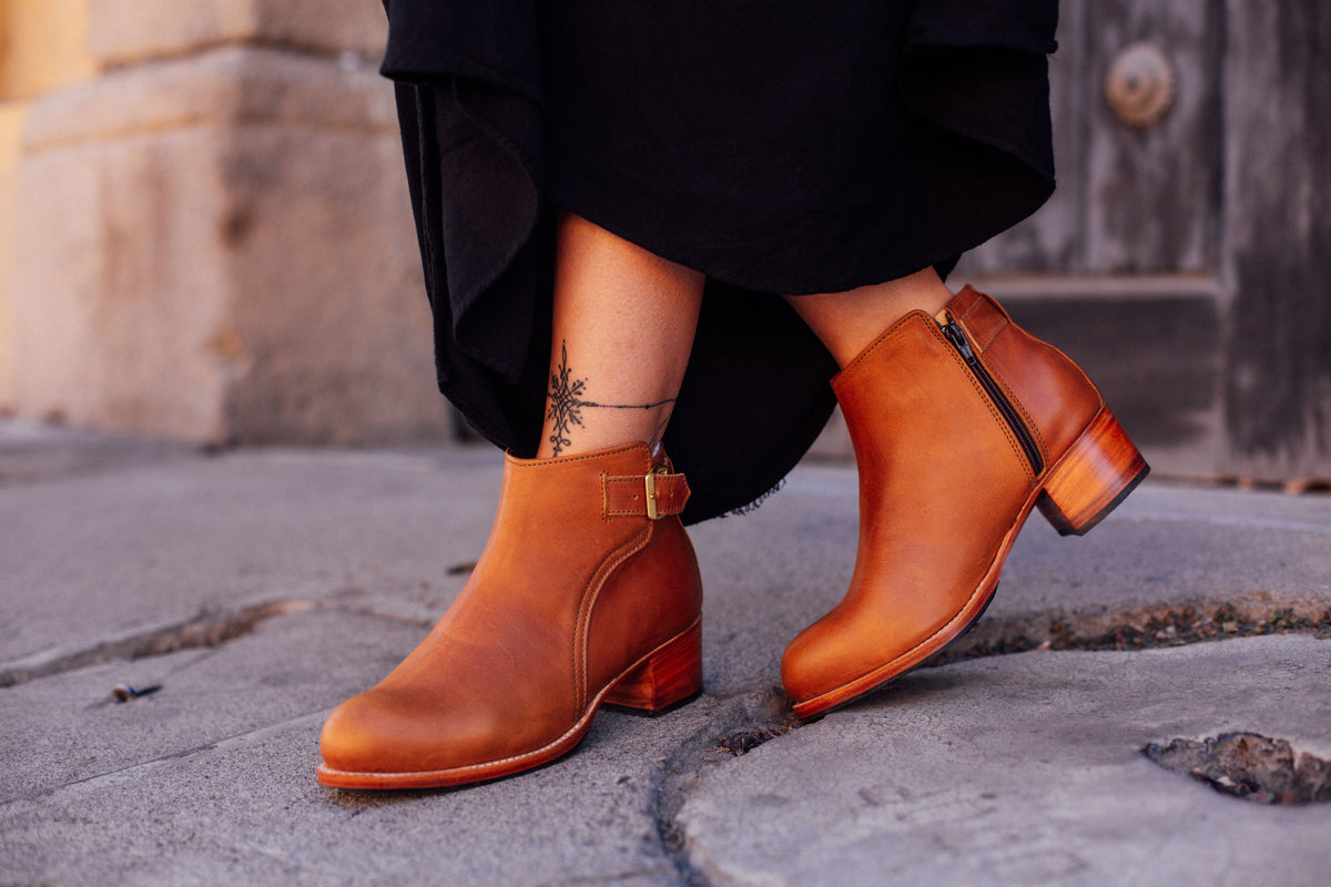 Women's tan buckle booties with block heels and side zippers paired with a black skirt