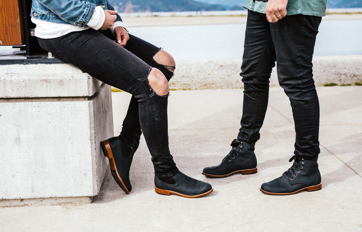 Black lace up ankle boots with brown soles worn outdoors by two people beside a concrete wall