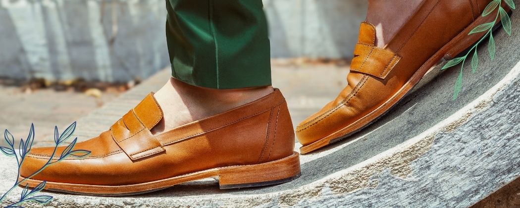 Handcrafted men's tan leather penny loafers worn with cuffed green pants on a stone walkway from the Adelante Made-to-Order Men's Shoes & Loafers Collection.
