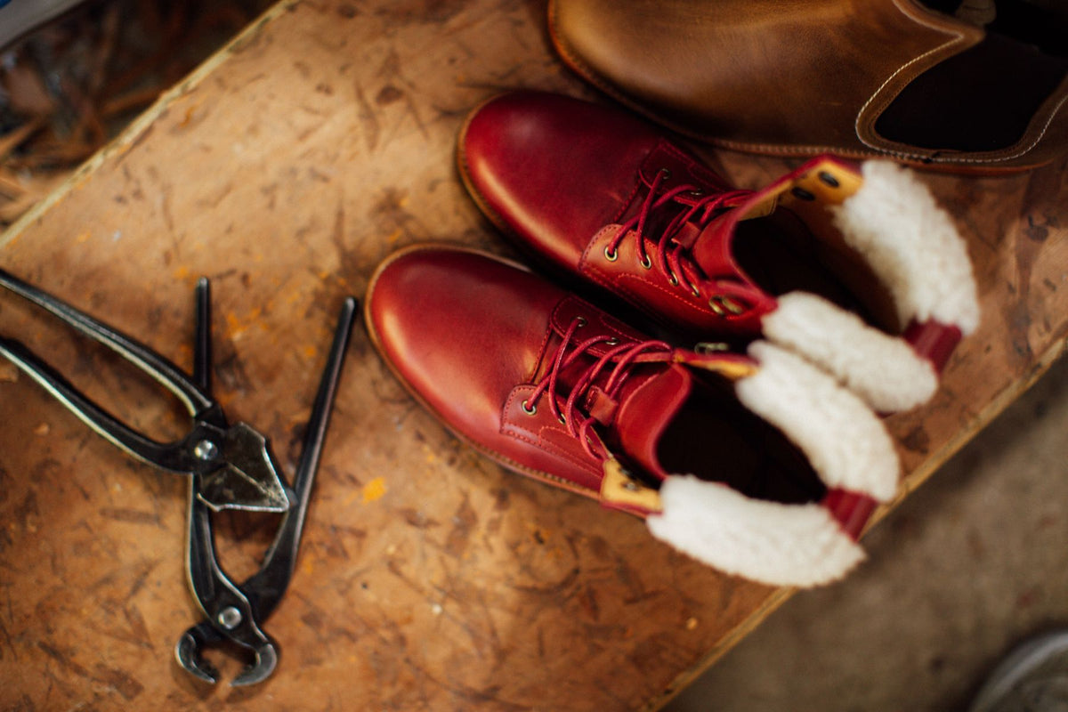 Handcrafted red leather lace-up boots with shearling lining on a cobbler's workbench beside shoemaker's pincers