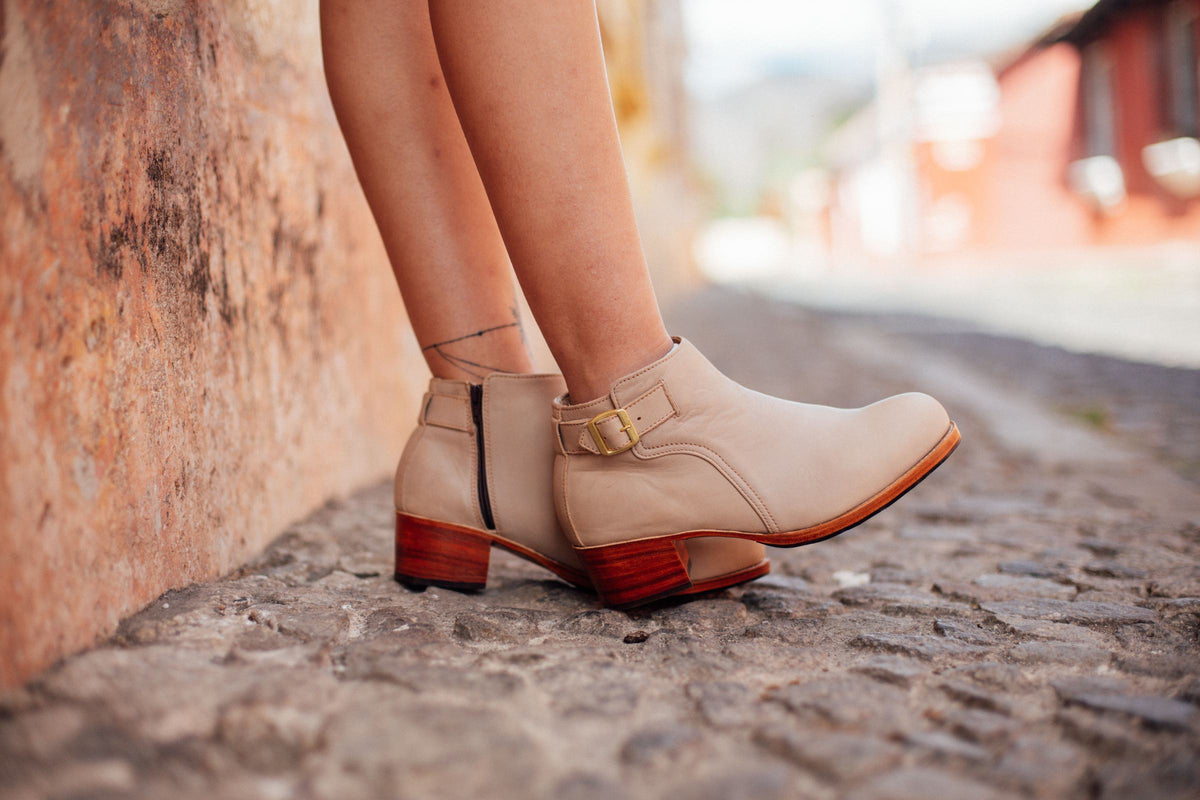 Women's cream leather ankle boots with a buckle strap, side zip and stacked block heels, worn on a cobblestone street beside an old plaster wall