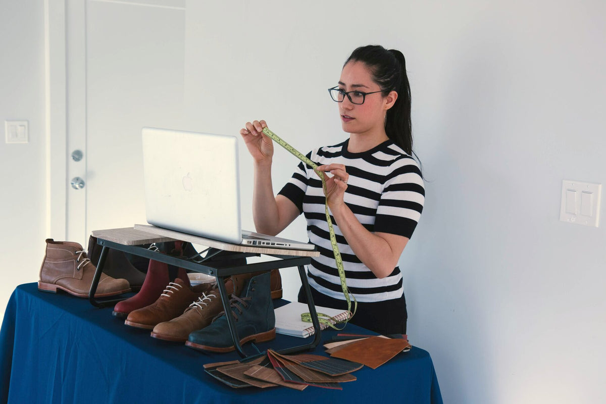 Woman holding a tape measure at a laptop during an Adelante virtual fitting, with leather boots and leather swatches on the table