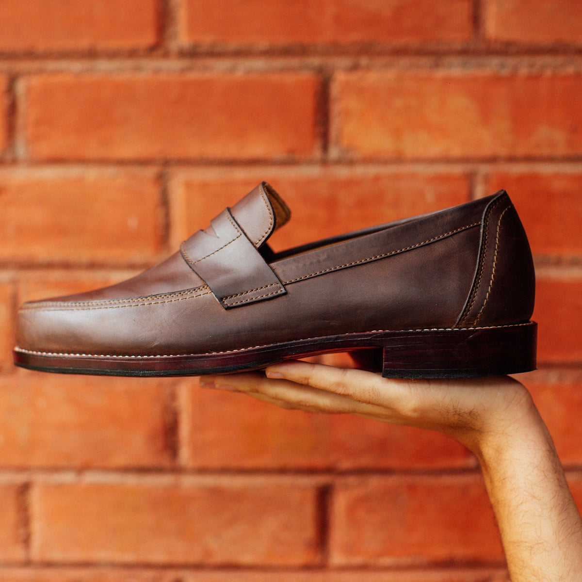 Adelante brown mens penny loafer held by hand, with a classic penny bridge, sleek toe, stitched detailing, and low stacked heel.